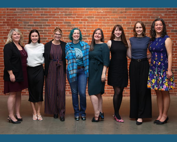 Image of 7 women standing in front of a brick wall smiling at the camera, professionally dressed.