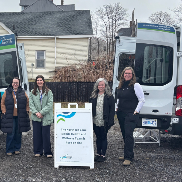 Four women in a parking lot stand behind a sandwich board sign that read "Northern Zone Mobile Health and Wellness Team" is here. Two vans are parked on both sides of the women. They wear jackets and sweaters. 