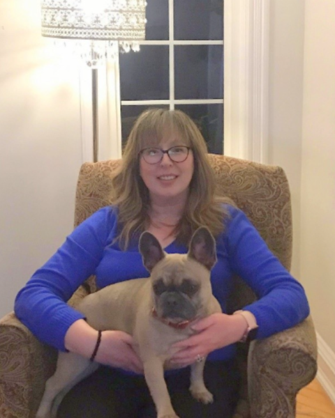 A woman sits in a chair inside wearing a blue shirt and is holding a pug dog.